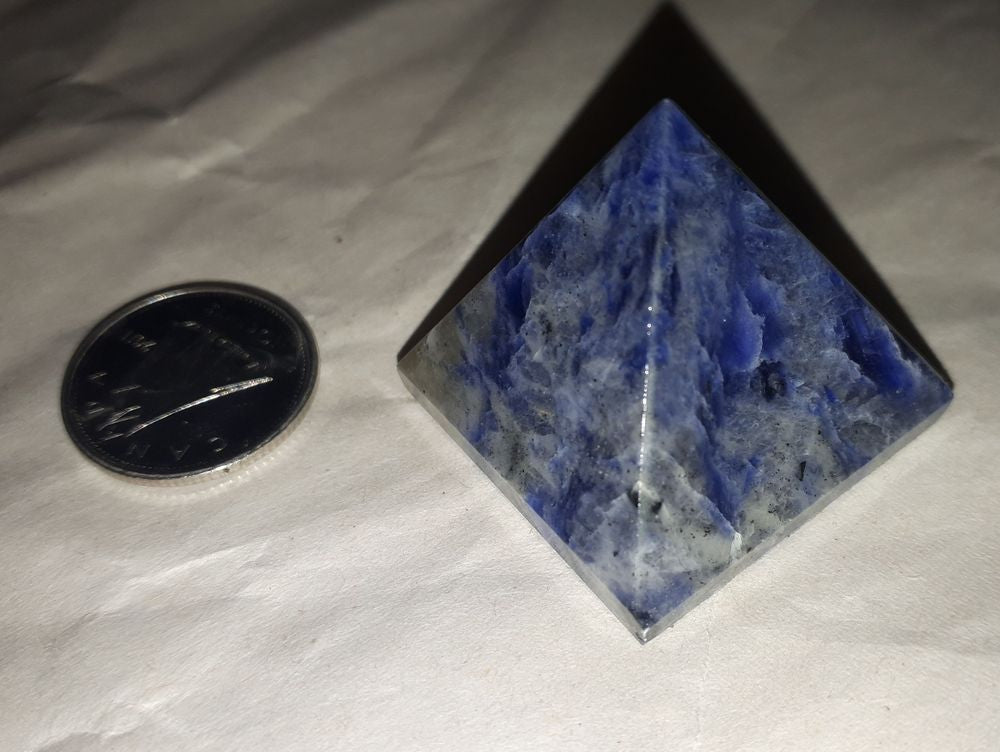 A natural sodalite pyramid of 25-28mm stone in blue colour with a silver coin on a serene white background.